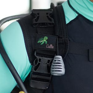 Op de afbeelding: Een zwarte schouderband voor een trimvest met een rechthoekige patch met een geborduurde groene schildpad en de naam Callie in wit schrift. De band bevat een zwarte plastic gesp en een grijze, getextureerde trekknop voor bediening.