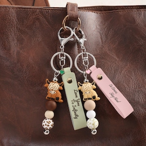 May include: Two keychains with Highland cow charms, attached to a brown leather bag. One keychain has a green tag with "Love You To Infinity", the other has a pink tag with "Be Brave, Stay Kind". Each keychain has a series of beads.