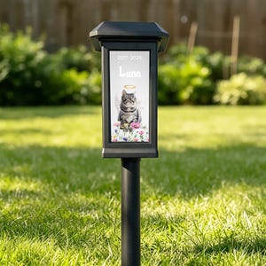 May include: Black solar-powered memorial garden light. The light features a framed image of a cat with angel wings and a halo, surrounded by flowers. The text "2017-2025" and "Luna" are displayed on the image. The light is set in a grassy yard.