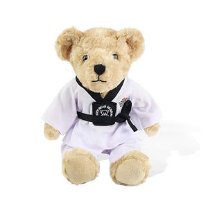 Puede incluir: Un oso de peluche vestido con un uniforme blanco de artes marciales con cinturón y ribetes negros. El uniforme lleva el texto "Eddy Bear Museum". El oso tiene pelaje marrón claro y está sentado.