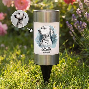 May include: A cylindrical solar garden light with a stainless steel top and base, featuring a printed illustration of a Dalmatian dog on a blue watercolor background, the name Bella, and the dates 2012-2026, with a small circular inset photo.