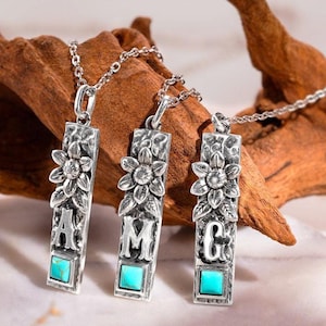 Puede incluir: Tres collares con colgantes plateados. Cada colgante tiene un diseño rectangular con un motivo floral, una letra y una piedra turquesa cuadrada. Las letras son A, M y G. Se muestran sobre un fondo de madera.