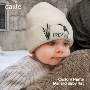 Puede incluir: Gorro de bebé color crema con un pato real bordado y el nombre "Grayson" en hilo verde. El gorro también presenta bordados de espadañas. La palabra "callie" está en la esquina superior izquierda. El texto "Custom Name Mallard Baby Hat" está en la parte inferior.