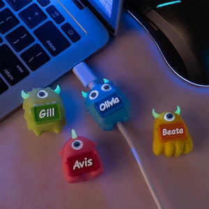 May include: A collection of colorful monster-shaped cable protectors. Each protector has a different color and a name on it: Gill, Olivia, Avis, and Beata. The protectors are near a laptop and a computer mouse.