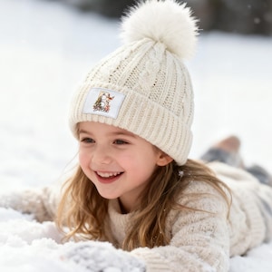 May include: Cream-colored cable knit beanie with a large white pom-pom. The beanie features a rectangular patch with the letter "B" and a Christmas tree design. The person in the image is wearing a matching cream-colored sweater.