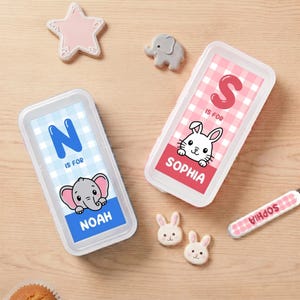 Może przedstawiać: Dwa spersonalizowane pojemniki na lunch z imionami "Noah" i "Sophia". Pojemnik "Noah" ma niebieski wzór w kratkę, grafikę słonia i literę "N". Pojemnik "Sophia" ma różowy wzór w kratkę, grafikę królika i literę "S".