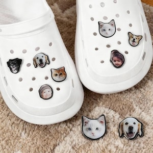 May include: White clogs decorated with animal-themed charms, featuring cats, dogs, and a baby's face. The charms are in various colours and styles, adding a playful touch to the footwear. The clogs are shown on a neutral-coloured rug.