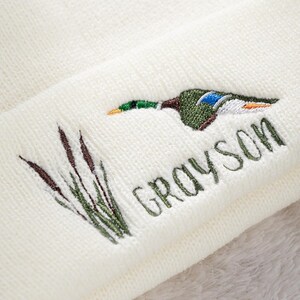 May include: Cream-colored knit beanie featuring an embroidered mallard duck in shades of green, blue, and brown, with the name "GRAYSON" in green script. The design includes embroidered cattails. A cozy and stylish winter accessory.