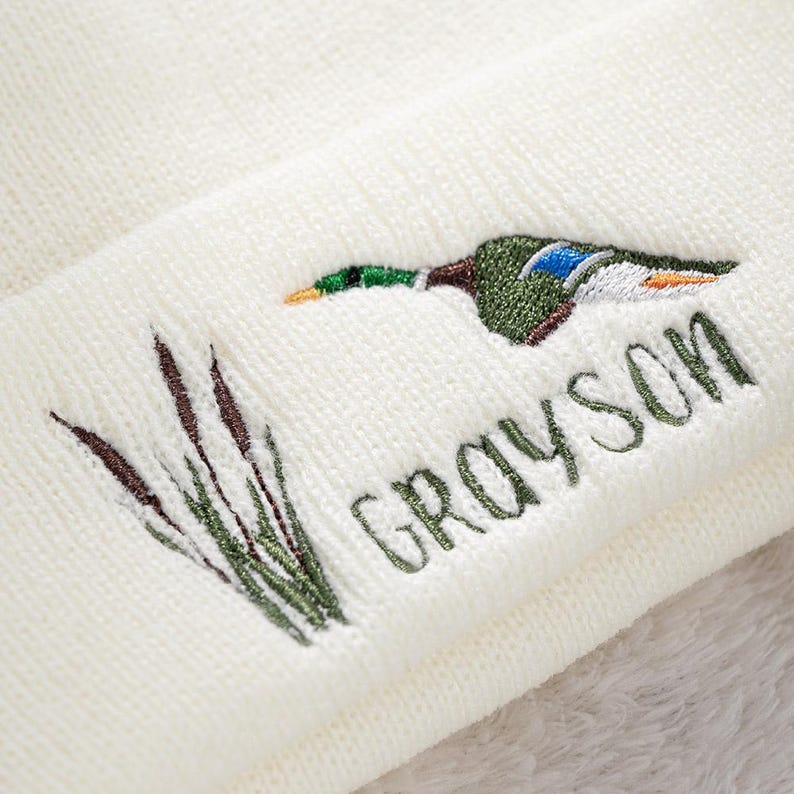 May include: Cream-colored knit beanie featuring an embroidered mallard duck in shades of green, blue, and brown, with the name "GRAYSON" in green script. The design includes embroidered cattails. A cozy and stylish winter accessory.