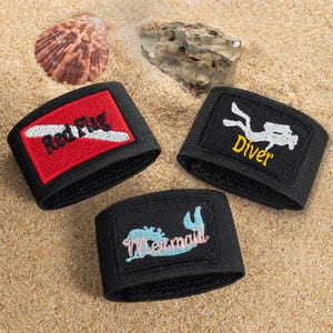 Puede incluir: Tres pulseras negras con parches bordados. Una tiene un diseño "Red Flag" rojo y blanco, otra presenta un buzo blanco y la palabra "Diver" en amarillo, y la última muestra un diseño "Mermaid" azul y rosa.