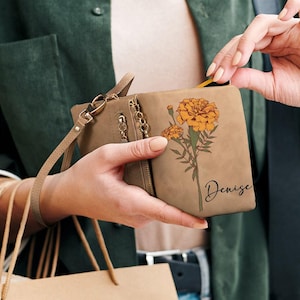K&ouml;nnte beinhalten: Eine hellbraune Geldb&ouml;rse mit einem Ringelblumen-Blumenmuster und dem Namen "Denise" in Schreibschrift. Die Geldb&ouml;rse hat eine Handschlaufe und mehrere Rei&szlig;verschl&uuml;sse. Die Ringelblumen sind orange und gelb mit gr&uuml;nen Stielen.