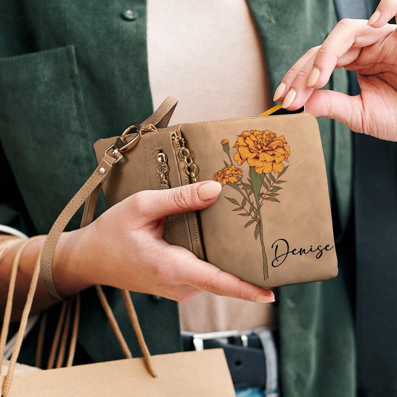 K&ouml;nnte beinhalten: Eine hellbraune Geldb&ouml;rse mit einem Ringelblumen-Blumenmuster und dem Namen "Denise" in Schreibschrift. Die Geldb&ouml;rse hat eine Handschlaufe und mehrere Rei&szlig;verschl&uuml;sse. Die Ringelblumen sind orange und gelb mit gr&uuml;nen Stielen.