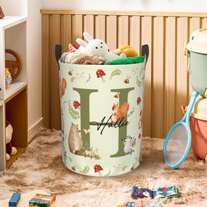May include: A cylindrical storage basket with black handles, filled with stuffed animals. The basket features a light green background with forest-themed illustrations, including a large letter "H" and the name "Hallie" in script.