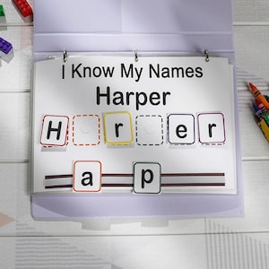 Może przedstawiać: Narzędzie edukacyjne z tekstem "I Know My Names Harper" wyświetlanym na białej kartce. Imię jest zapisane za pomocą kolorowych kafelków z literami. Segregator jest fioletowy i leży na białej drewnianej powierzchni.