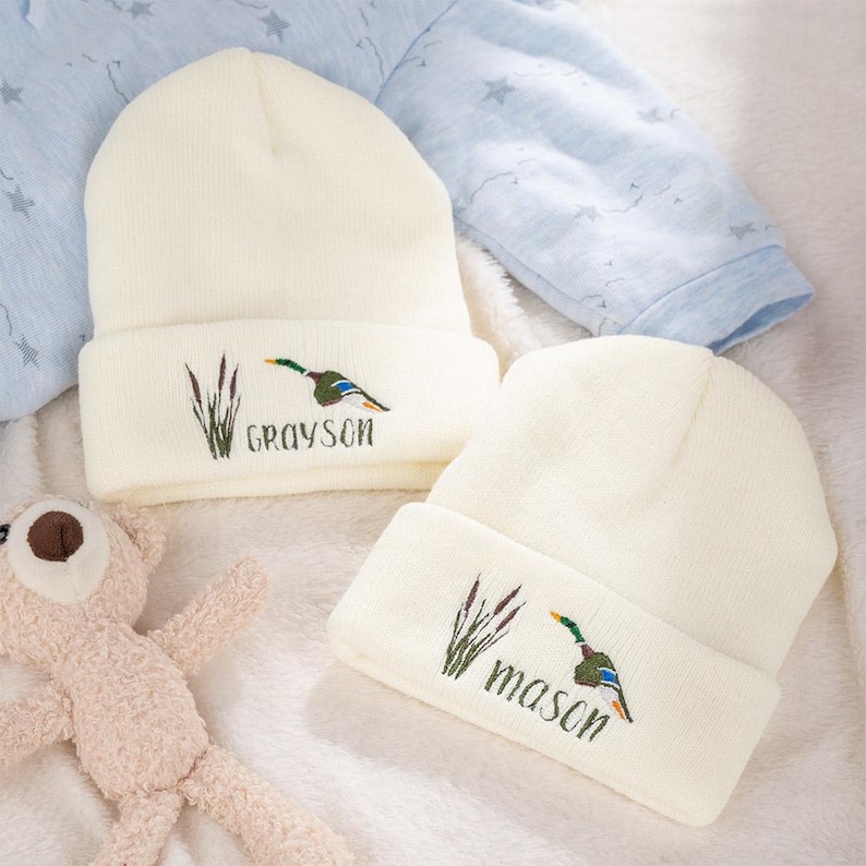 May include: Two cream-colored knit beanies with embroidered duck and cattail designs. One beanie says "Grayson" and the other says "Mason". A light blue baby outfit with star patterns is in the background.