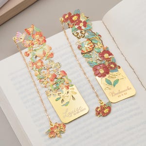 May include: Two gold-toned metal bookmarks featuring intricate cut-out floral and butterfly designs with enamel accents in red, pink, and green. One bookmark is engraved with the name Louise and a date, while the other features the text Bookmarks 2025.