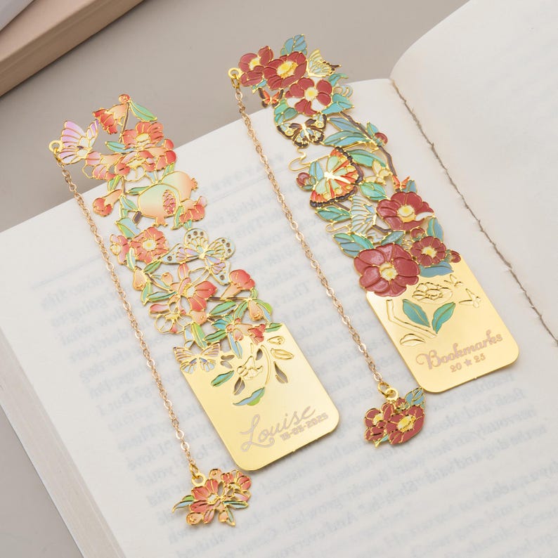 May include: Two gold-toned metal bookmarks featuring intricate cut-out floral and butterfly designs with enamel accents in red, pink, and green. One bookmark is engraved with the name Louise and a date, while the other features the text Bookmarks 2025.