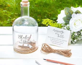 May include: A clear glass bottle with a cork stopper, containing a rolled-up message and twine. The bottle has the names "Blake & Mary" printed on it. A card with the text "message in a bottle" and a pencil are also present.