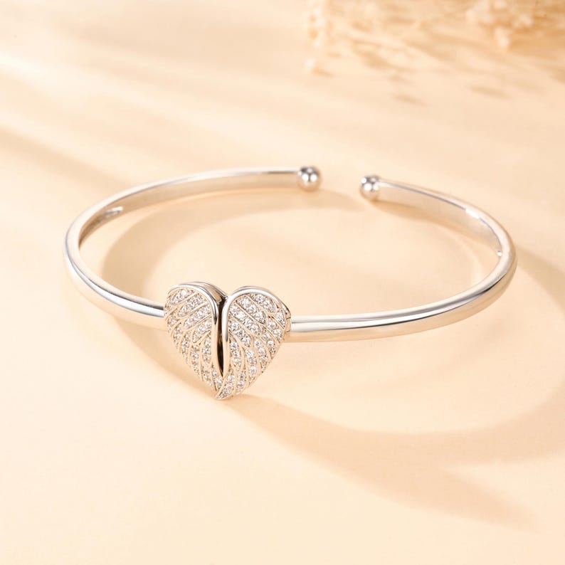 May include: A silver-toned cuff bracelet with a heart-shaped charm. The charm is encrusted with small, sparkling crystals, and the bracelet has a smooth, polished finish. The bracelet is a delicate piece of jewelry.