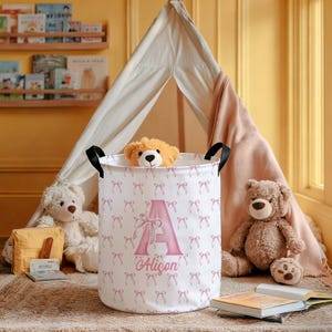 May include: A white laundry hamper with a pink bow pattern and black handles. The hamper features a large pink letter "A" and the name "Alison". A teddy bear peeks over the top. Other teddy bears and books are nearby.