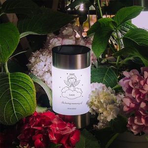 Peut inclure: Lampe solaire commémorative cylindrique avec finition en acier inoxydable et dessus noir. Elle présente un motif gravé d'un ange sur un nuage, avec le nom "Leon" et l'inscription "In loving memory of 10.14.2023".