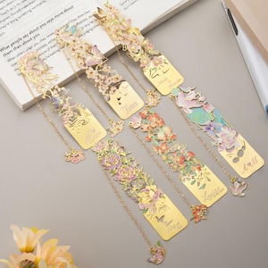 May include: Five gold-toned metal bookmarks featuring intricate cut-out designs of flowers, butterflies, and leaves with pastel enamel accents. Each bookmark includes a delicate gold chain with a small floral charm and personalized engraved names.