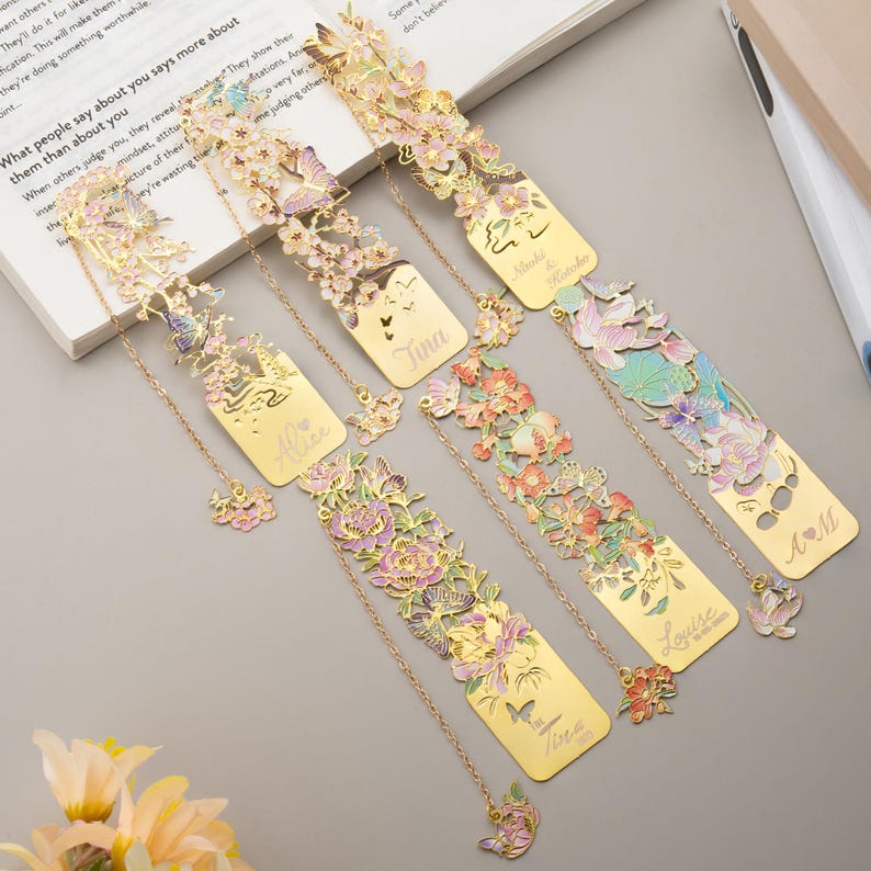 May include: Five gold-toned metal bookmarks featuring intricate cut-out designs of flowers, butterflies, and leaves with pastel enamel accents. Each bookmark includes a delicate gold chain with a small floral charm and personalized engraved names.