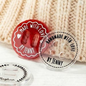 May include: Close-up of three decorative buttons. One is red with the text "MADE WITH LOVE BY JENNY". Another is clear with the text "HANDMADE WITH LOVE BY JENNY". The third is clear with a floral design and the name "Meghan".