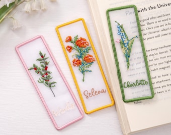 May include: Three rectangular bookmarks made of sheer white fabric with embroidered floral designs and names. The left bookmark has red berries, the center has orange flowers labeled Selena, and the right has blue flowers labeled Charlotte.