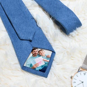 Może przedstawiać: Niebieski dżinsowy krawat ze zdjęciem mężczyzny i dziecka trzymających kartkę z napisem "I ❤️ Daddy". Krawat leży na białej, puszystej powierzchni. W tle srebrny zegarek z białą tarczą.