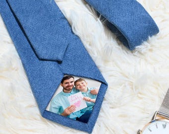 Puede incluir: Una corbata de mezclilla azul con una foto de un hombre y un niño sosteniendo una tarjeta que dice "I ❤️ Daddy". La corbata está sobre una superficie blanca y esponjosa. Un reloj plateado con esfera blanca en el fondo.