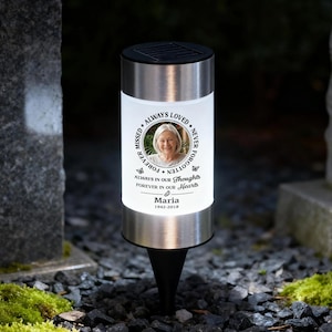 Puede incluir: Una lámpara solar conmemorativa cilíndrica con tapa y base de metal plateado. El centro blanco presenta un retrato circular de una mujer mayor, rodeado por el texto ALWAYS LOVED, NEVER FORGOTTEN, FOREVER MISSED y el nombre Maria 1942-2018.