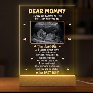 May include: A glowing acrylic plaque with the words "DEAR MOMMY" and a sonogram image. The plaque features a poem about a baby's love, set on a wooden base. The overall theme is pregnancy and baby showers.