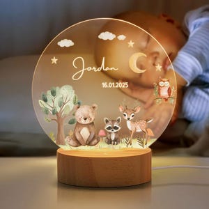 Puede incluir: Luz de noche personalizada con una escena del bosque, con un oso, un mapache y un ciervo. El disco acrílico transparente tiene el nombre "Jordan" y la fecha 16.01.2025. Se apoya en una base de madera y emite una luz cálida.