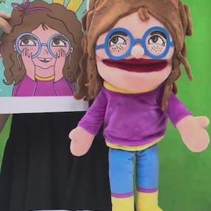 May include: A soft puppet with brown hair, blue-rimmed spectacles, and a purple bow. The puppet is dressed in a purple top, blue trousers, and yellow boots with blue toes. A drawing of a girl with glasses is held beside the puppet.