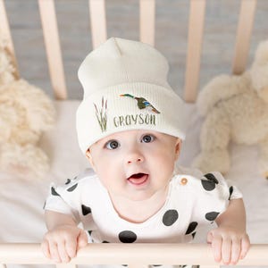 May include: A cream-colored baby beanie with an embroidered duck and cattails design. The name "Grayson" is embroidered below the design. The baby is wearing a black and white polka dot onesie and is in a crib.