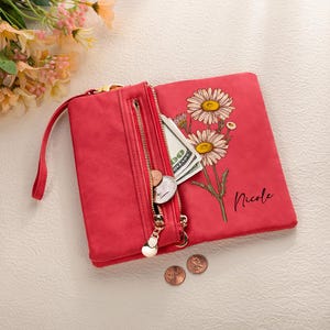 K&ouml;nnte beinhalten: Rotes Portemonnaie mit goldenem Rei&szlig;verschluss, Handschlaufe und G&auml;nsebl&uuml;mchen-Blumendesign. Das Portemonnaie ist ge&ouml;ffnet und zeigt Bargeld und M&uuml;nzen. Der Name "Nicole" ist aufgedruckt.