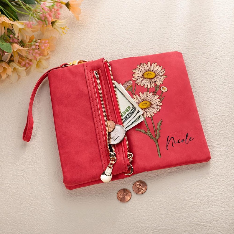 K&ouml;nnte beinhalten: Rotes Portemonnaie mit goldenem Rei&szlig;verschluss, Handschlaufe und G&auml;nsebl&uuml;mchen-Blumendesign. Das Portemonnaie ist ge&ouml;ffnet und zeigt Bargeld und M&uuml;nzen. Der Name "Nicole" ist aufgedruckt.