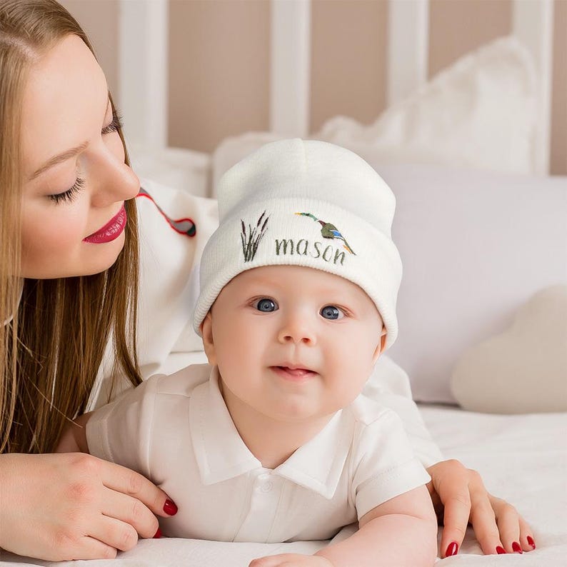 May include: A white baby beanie with a colorful bird and cattail design, and the name "mason" printed in a script font. The baby is wearing a white shirt and is lying on a white surface.