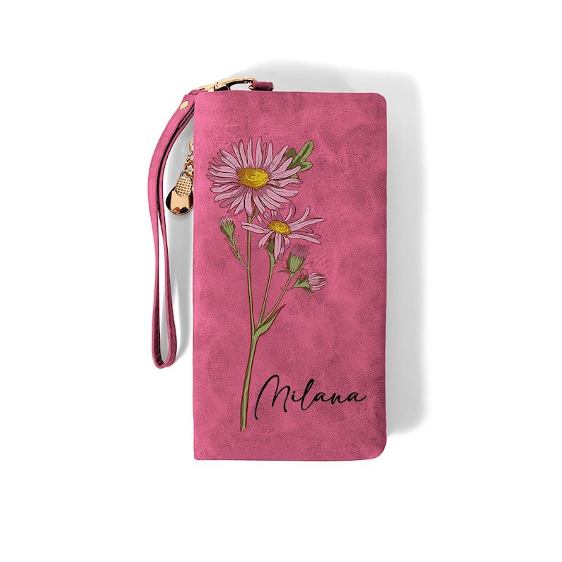 K&ouml;nnte beinhalten: Eine pinkfarbene Geldb&ouml;rse mit floralem Design und dem Namen "Milana" in Schreibschrift. Die Geldb&ouml;rse hat eine Handschlaufe und einen goldfarbenen Rei&szlig;verschluss. Das florale Design umfasst rosa Bl&uuml;ten mit gelben Zentren.