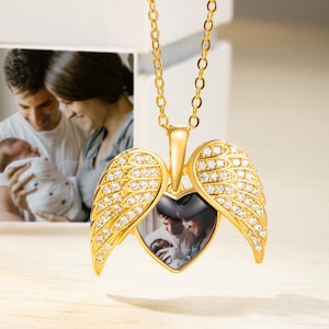 Può includere: Una collana con pendente a forma di cuore dorato con ali d'angelo, incastonate con piccole pietre trasparenti. Il pendente si apre per rivelare una piccola fotografia. La collana è esposta davanti a una fotografia incorniciata di una famiglia.