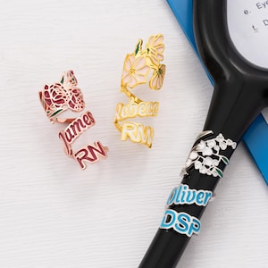 Puede incluir: Tres anillos con temática floral con nombres e iniciales personalizados. Uno es de oro rosa con una flor roja y los nombres "James" y "RN". Otro es dorado con una flor blanca y los nombres "Rebecca" y "RM". El tercero es plateado con los nombres "Oliver" y "DSP".