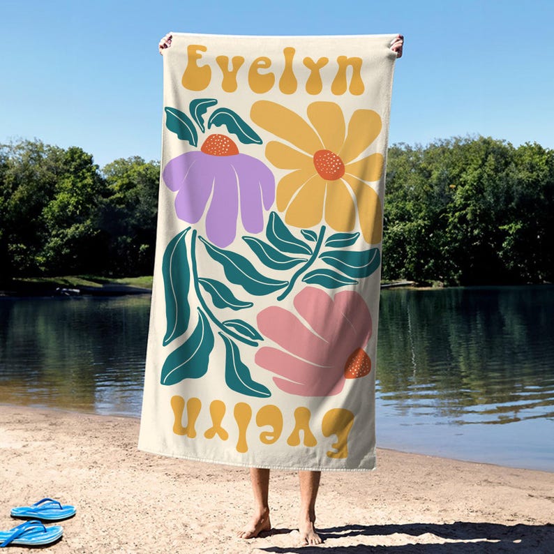May include: A cream-colored beach towel featuring a floral design with a purple, yellow, and pink flowers, and teal leaves. The name "Evelyn" is printed in a retro font at the top and bottom of the towel. The towel is held up on a beach.