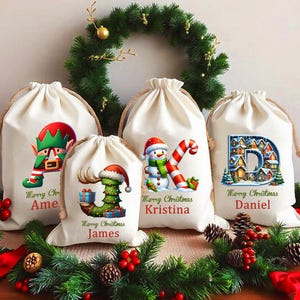 May include: Five cream-coloured drawstring Christmas gift bags with festive designs. Each bag features a unique holiday graphic, including an elf, a Christmas tree, a snowman, and a letter "D" with a village scene. The bags are decorated with the names "Ame", "James", "Kristina", and "Daniel".