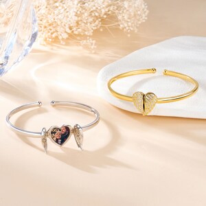 May include: Two open-ended bracelets. One is silver with a heart-shaped locket that opens to reveal a photo, and small wing charms. The other is gold, with a heart-shaped charm adorned with small, sparkling stones.