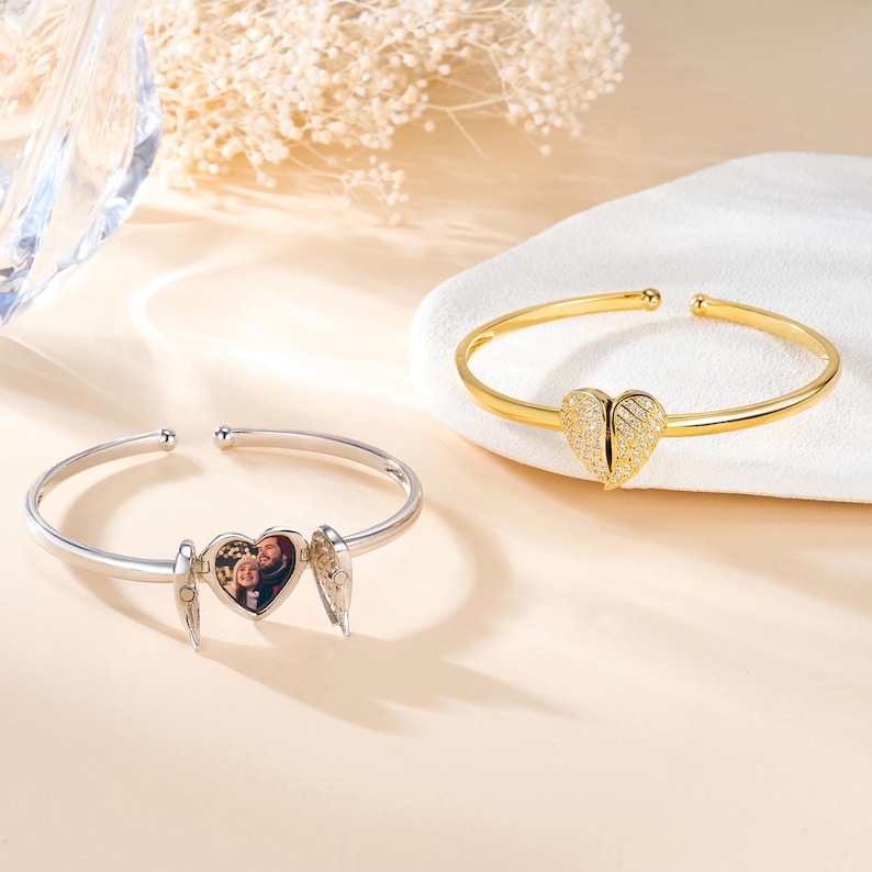 May include: Two open-ended bracelets. One is silver with a heart-shaped locket that opens to reveal a photo, and small wing charms. The other is gold, with a heart-shaped charm adorned with small, sparkling stones.