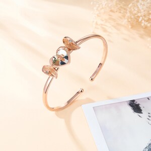 May include: Rose gold cuff bracelet featuring a heart-shaped charm with a photo and two decorative wing accents. The open design allows for easy wear. The bracelet is a stylish accessory.