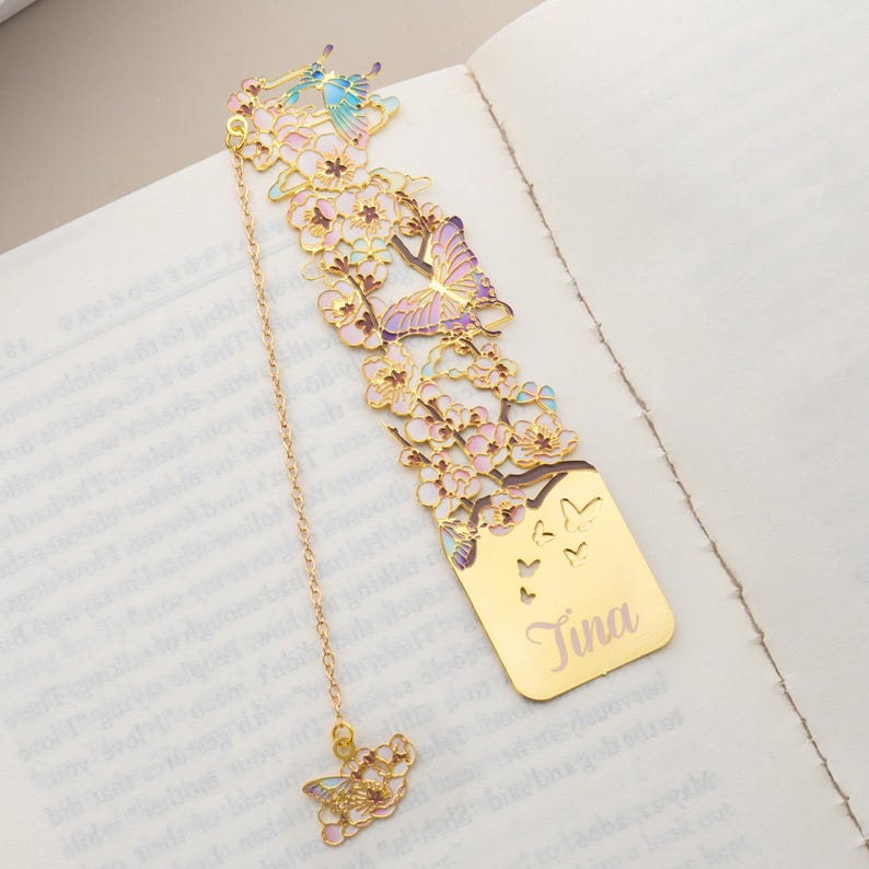 May include: A gold-toned metal bookmark featuring an intricate cutout design of cherry blossoms and butterflies with pastel enamel accents. The bottom section is a solid gold-toned plate engraved with the name Tina, attached to a chain with a charm.