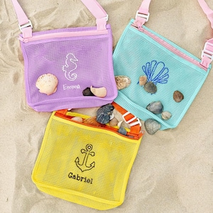 Könnte beinhalten: Drei bunte Strandtaschen aus Netzstoff, gefüllt mit Muscheln. Die Taschen sind lila, türkis und gelb, jede mit einem anderen gestickten Design: einem Seepferdchen, einer Muschel und einem Anker. Die Taschen haben rosa Träger und Reißverschlüsse, und die Namen "Emma" und "Gabriel" sind auf die Taschen gestickt.