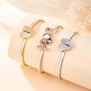 May include: Three heart-shaped bracelets in gold, rose gold, and silver. Each bracelet features a heart design with wing accents. The gold bracelet has a textured heart, the rose gold has a photo, and the silver has a crystal-studded heart.
